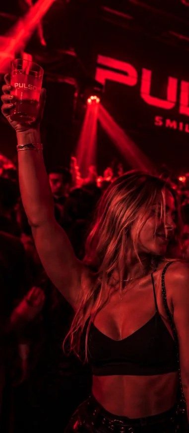 Una chica levanta su vaso de PULSO bajo las luces rojas
